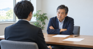 腕を組んで候補者を見つめる経営者と、緊張して座る面接の候補者